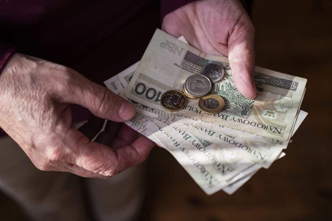 Dłoń starszej osoby trzymająca banknoty i monety na tle ciemnego tła.