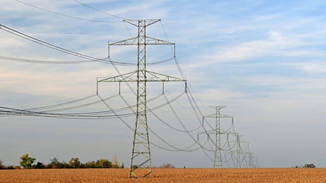 Widok na linie wysokiego napięcia na polu z drzewami w tle, pod niebieskim niebem z chmurami.