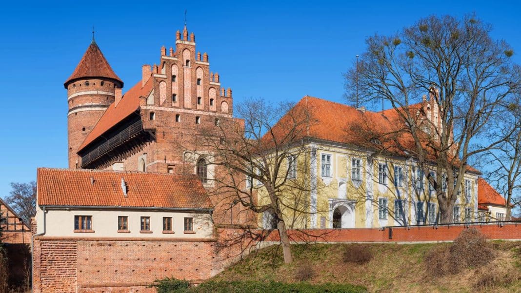 Zabytkowy zamek na Warmii i Mazurach z czerwonym dachem i wieżą, otoczony drzewami, na tle błękitnego nieba.