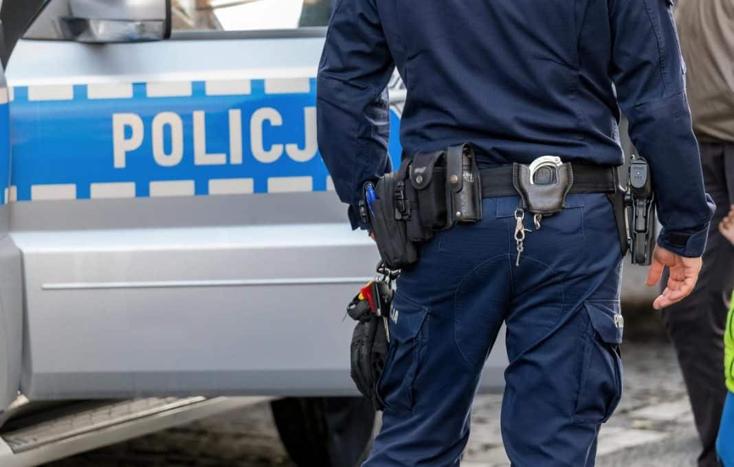 Policjant w mundurze z widocznym sprzętem na pasie, obok radiow&oacute;z z napisem 'Policja'.