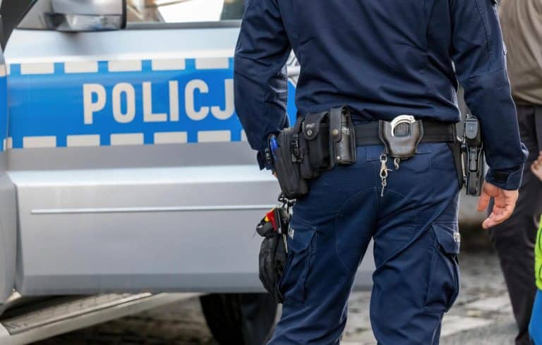Policjant w mundurze z widocznym sprzętem na pasie, obok radiow&oacute;z z napisem 'Policja'.