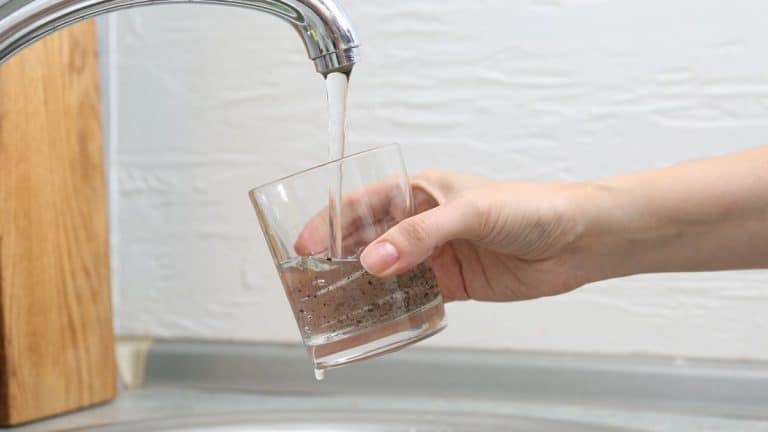 Kobieta nalewa wodę z kranu do szklanki, w tle biała ściana.