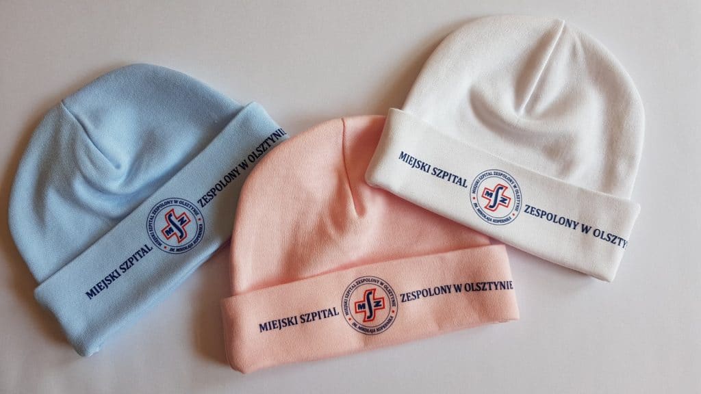 Trzy beanie z logo Miejskiego Szpitala w Olsztynie w r&oacute;żnych kolorach: niebieskim, r&oacute;żowym i białym.