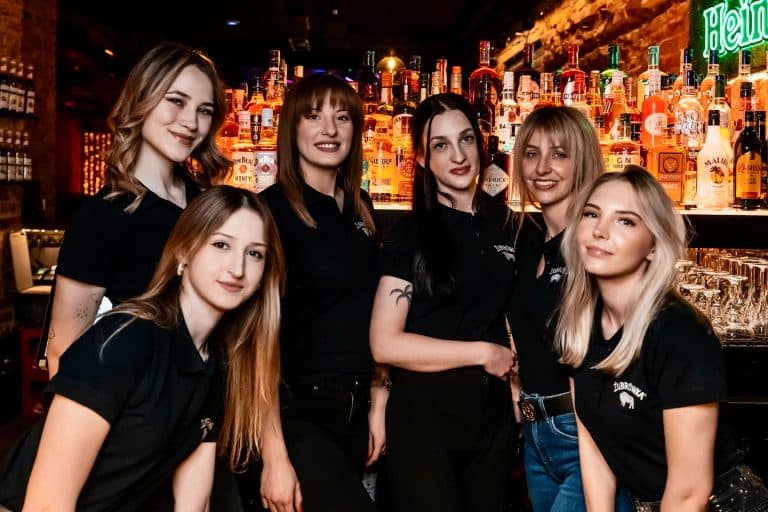 Grupa kobiet stojących za barem w nowym klubie na star&oacute;wce Olsztyna, w tle kolorowe butelki alkoholu.