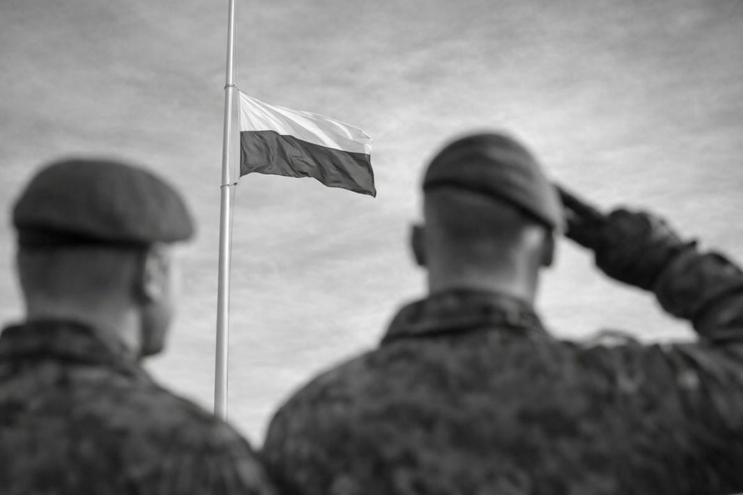 Dw&oacute;ch żołnierzy salutuje przy fladze Polski na tle nieba, w czerni i bieli.
