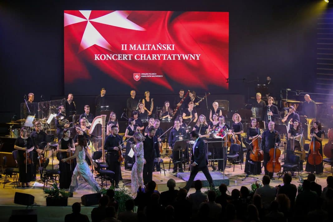 Orkiestra symfoniczna na scenie podczas koncertu charytatywnego w Olsztynie, na dużym ekranie napis o wydarzeniu.