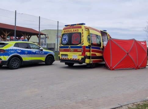 Na zdjęciu karetka i policja na ulicy po tragicznej zdarzeniu, w tle ogrodzenie i drzewa.
