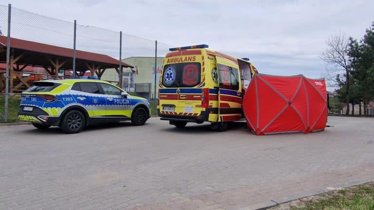 Na zdjęciu karetka i policja na ulicy po tragicznej zdarzeniu, w tle ogrodzenie i drzewa.