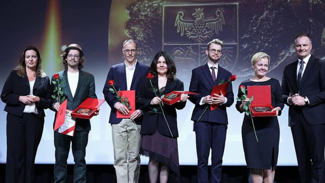 Grupa os&oacute;b na scenie podczas gali, trzymająca nagrody i kwiaty, w tle herb wojew&oacute;dztwa warmińsko-mazurskiego.