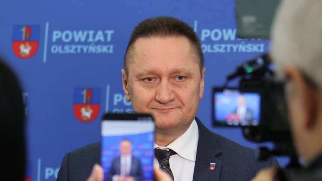 Starosta Olsztyna podczas wywiadu, w tle logo powiatu olsztyńskiego.