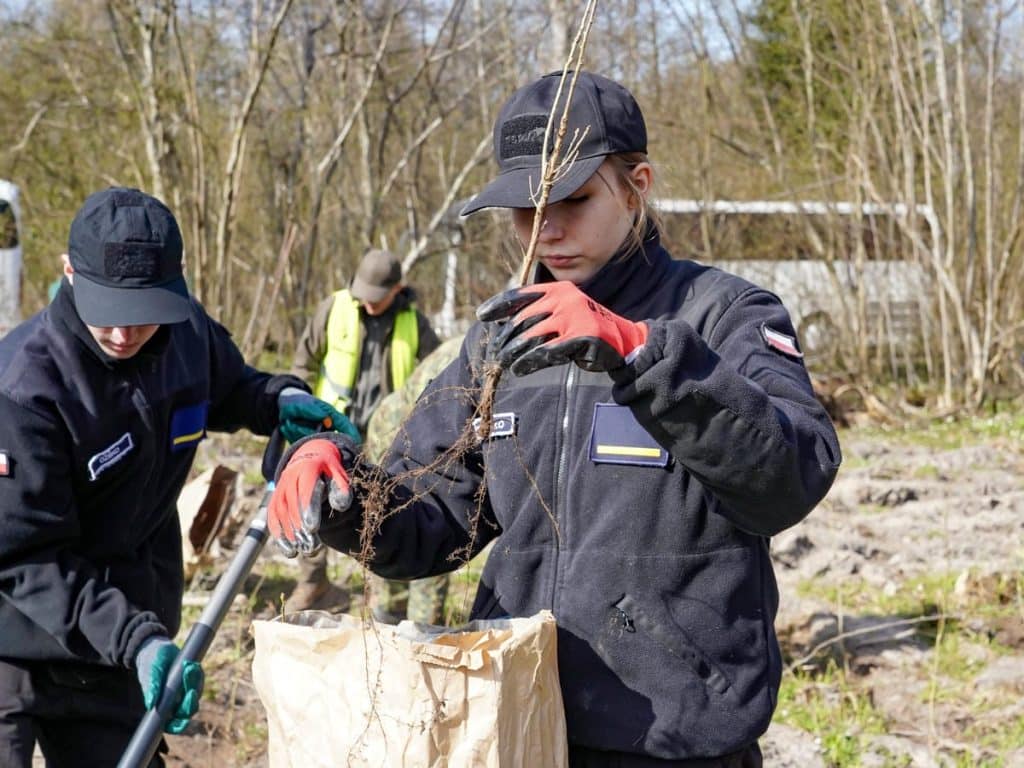 Młodzież sadzi drzewa podczas akcji w Wilczym Szańcu na Mazurach, w tle las i wiosenna przyroda.