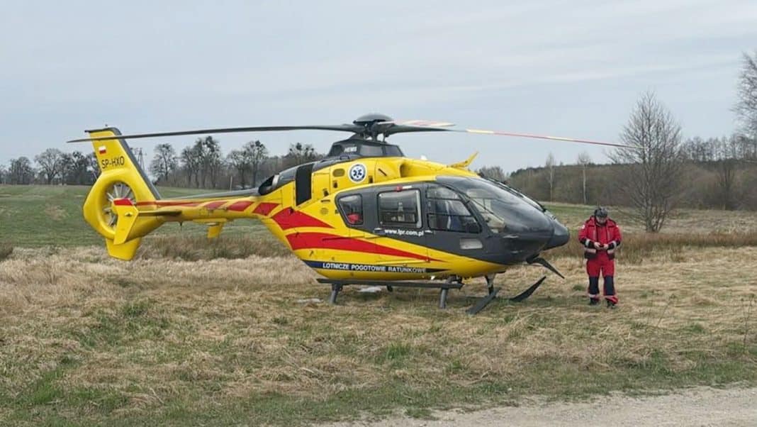 Ż&oacute;łty helikopter ratunkowy i ratownik na polu, w tle drzewa i niebo.