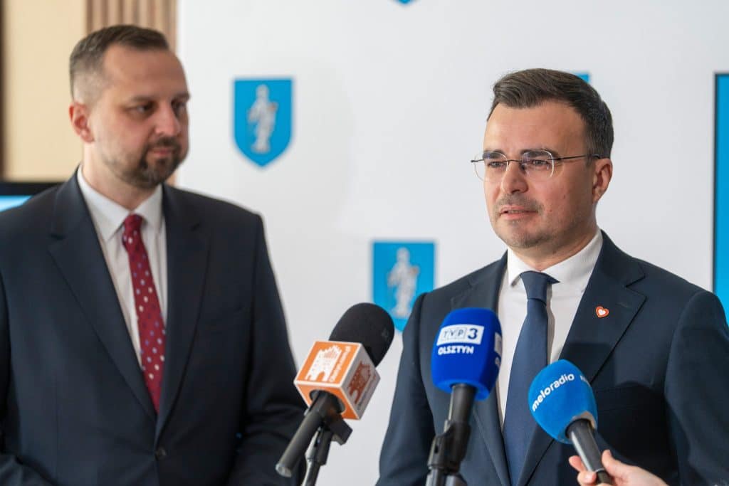 Dw&oacute;ch mężczyzn w garniturach podczas konferencji prasowej, z mikrofonami i logo miasta w tle.