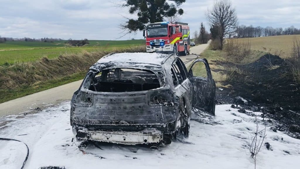 Spalony samoch&oacute;d na poboczu drogi po pożarze, w tle straż pożarna.