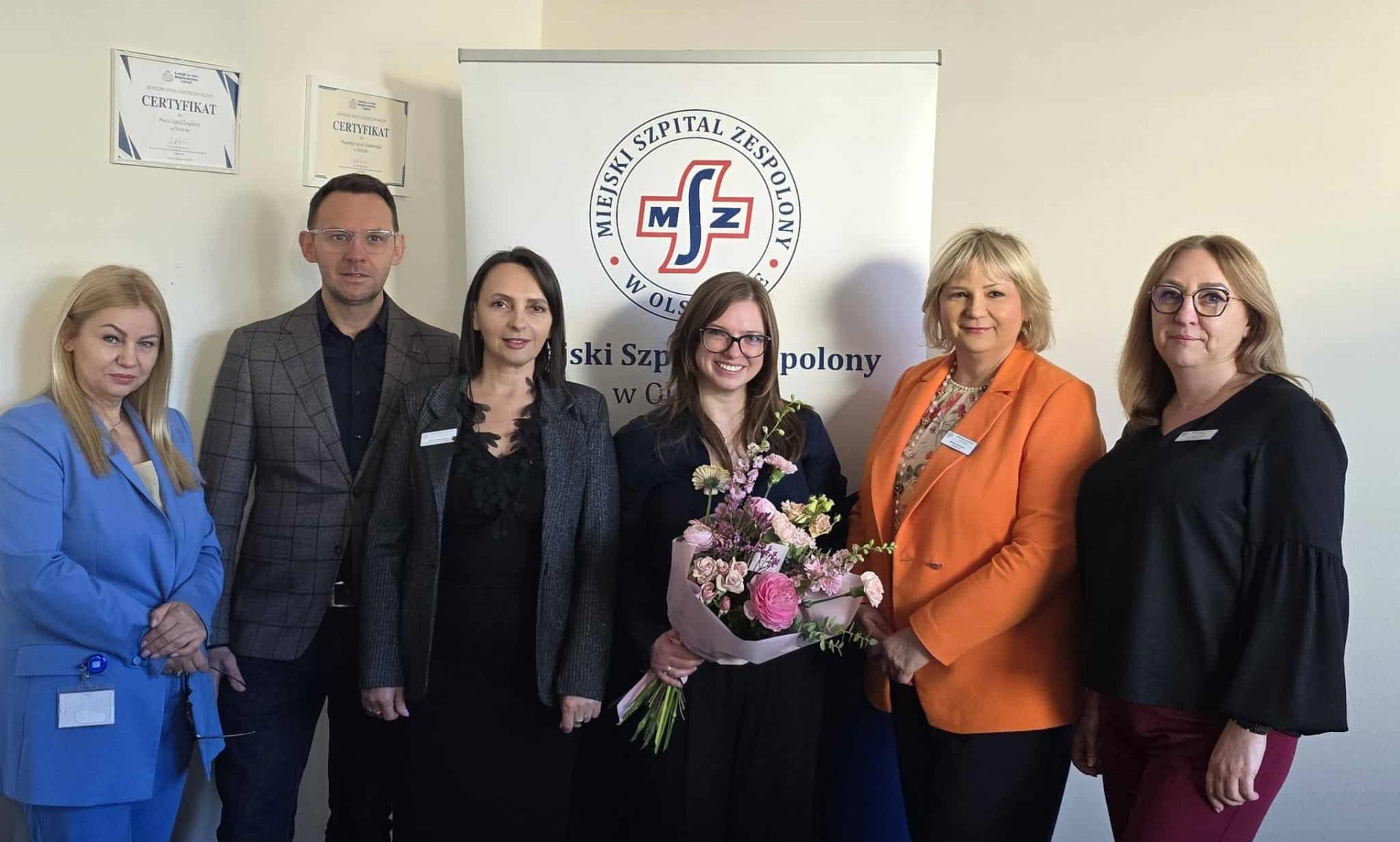 Grupa os&oacute;b na tle logo szpitala, jedna kobieta trzyma bukiet kwiat&oacute;w.