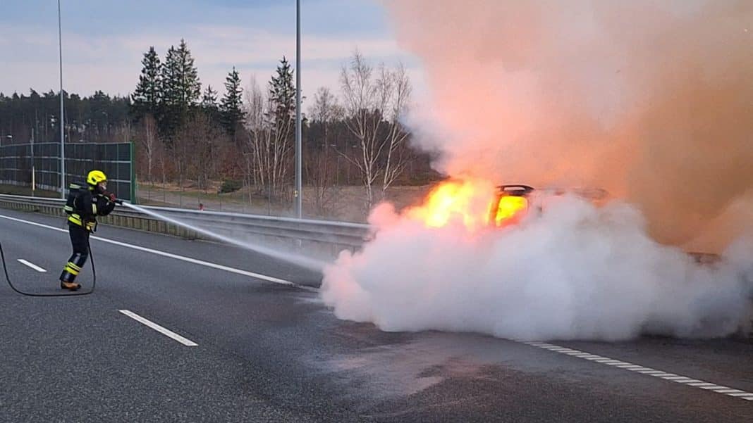 Strażak gasi pożar samochodu na trasie S7, w tle dym i płomienie, widok na okoliczne drzewa.