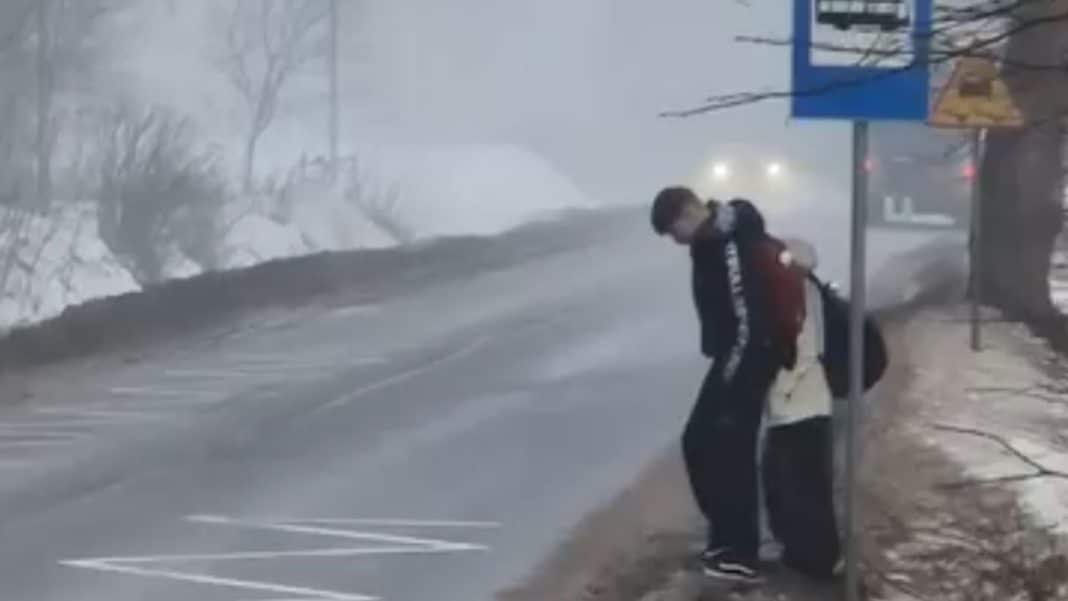 Dzieci czekające na autobus szkolny na krawędzi głębokiego rowu przy ruchliwej drodze, stojąc za metalowym słupkiem.