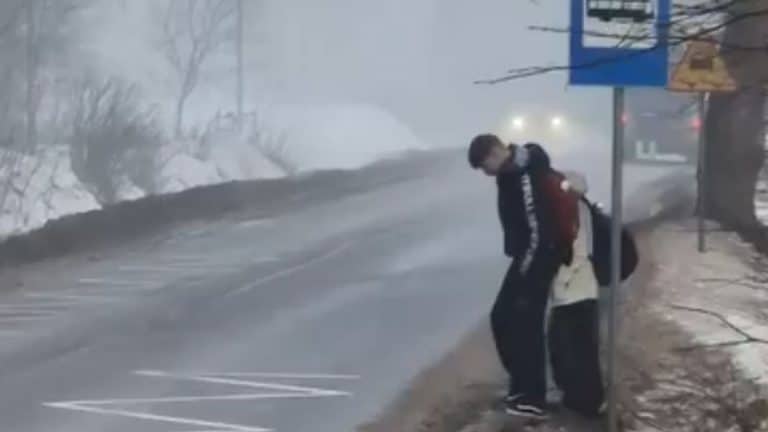 Dzieci czekające na autobus szkolny na krawędzi głębokiego rowu przy ruchliwej drodze, stojąc za metalowym słupkiem.