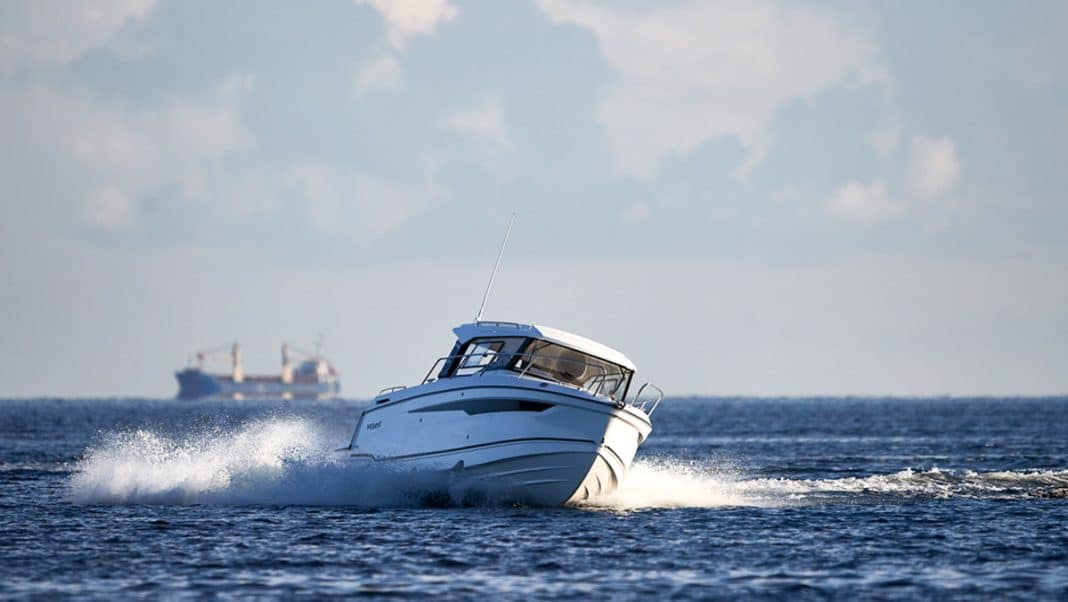 Biała ł&oacute;dź motorowa płynie po wodzie, za nią widoczny statek na horyzoncie.