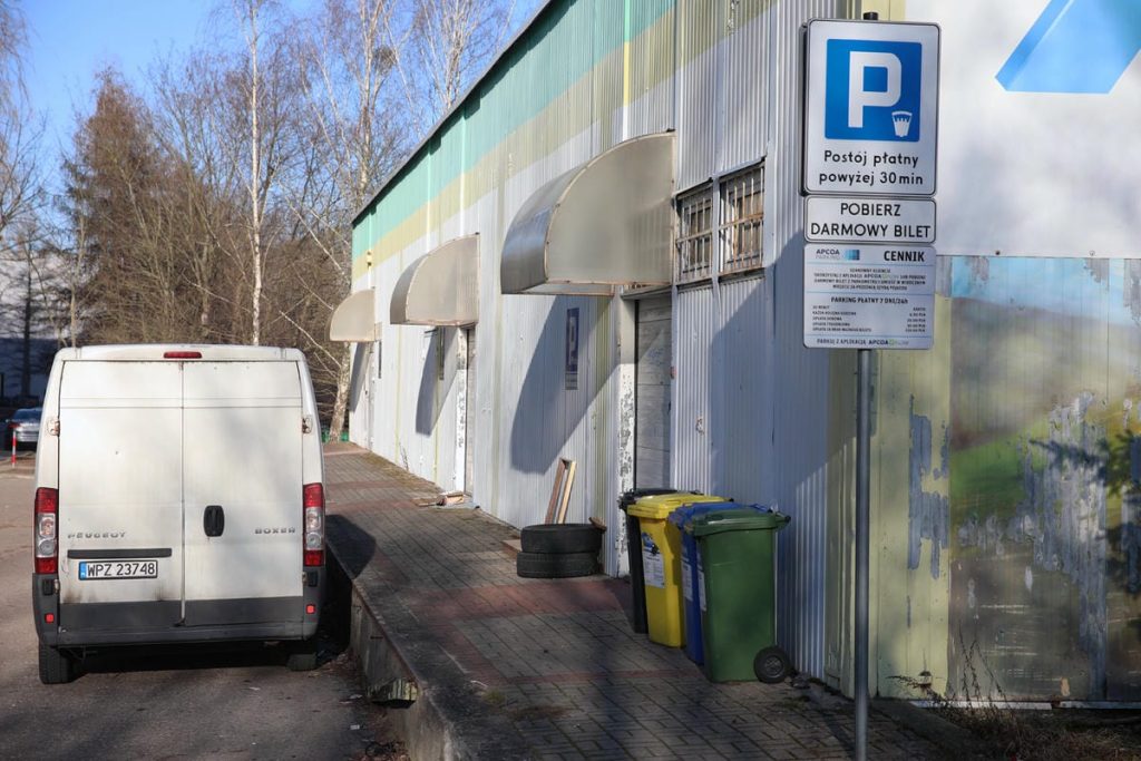 Budynek handlowy z odpadkami i parkingiem, w tle drzewa i niebo.