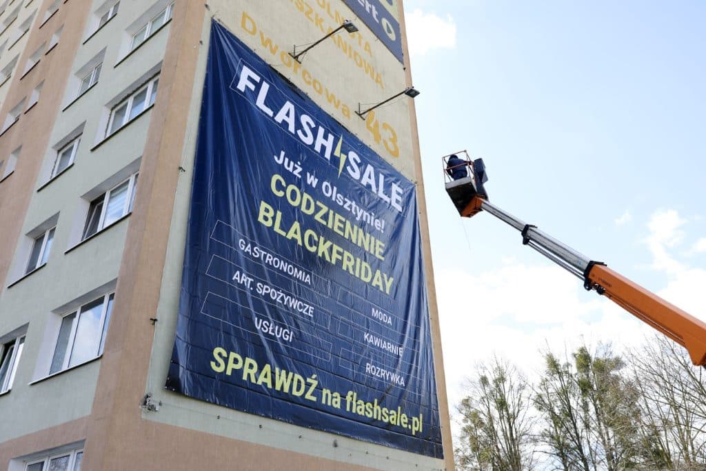 Pracownik na podnośniku montuje baner reklamowy na budynku w Olsztynie.