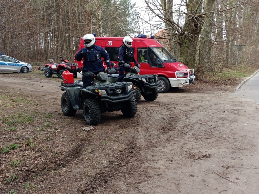 Na miejscu akcji służb ratunkowych w lesie, widoczne policyjne pojazdy i funkcjonariusze na motocyklach i quadach.