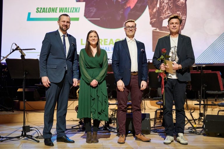 Grupa czterech os&oacute;b na scenie podczas uroczystości, jedna z nich trzyma r&oacute;żę.