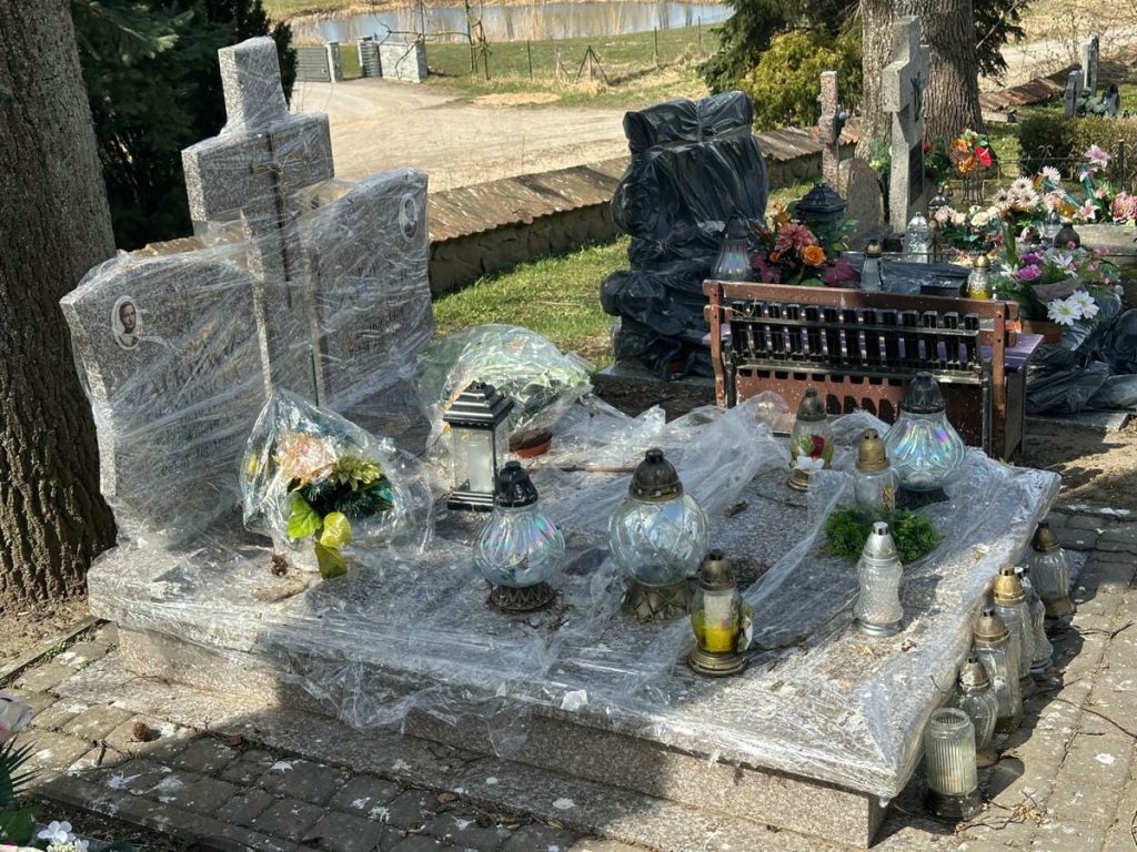 Nagrobek owinięty folią, otoczony lampami i kwiatami, w tle drzewa i cmentarz.