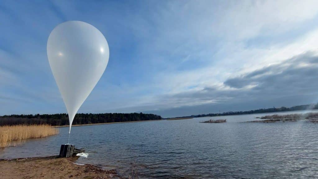 Balon na wodzie przy brzegu jeziora, w tle chmury i las, na zdjęciu widoczny balon z przyczepionym obiektem.
