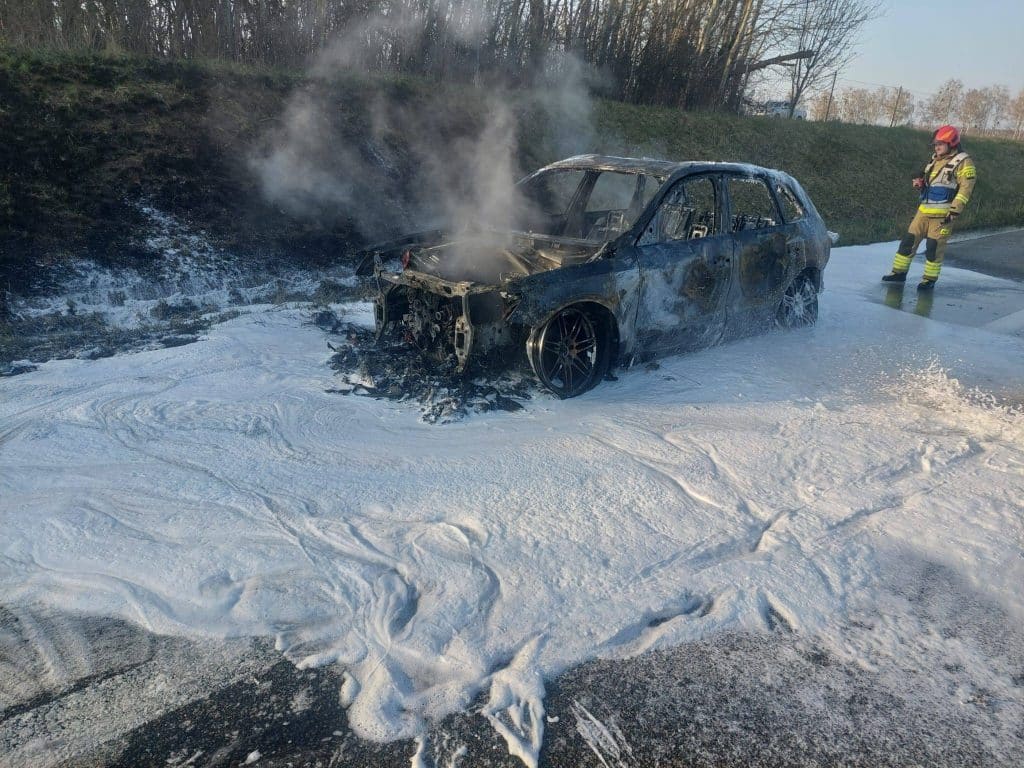 Spalony samoch&oacute;d marki Audi na drodze ekspresowej, z widocznym dymem i strażakiem obok.