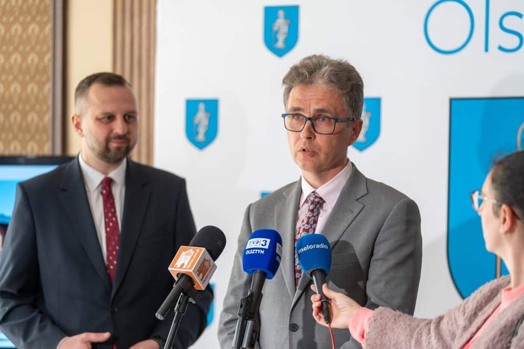 Mężczyzna w okularach i garniturze przemawia do medi&oacute;w podczas konferencji w Olsztynie.