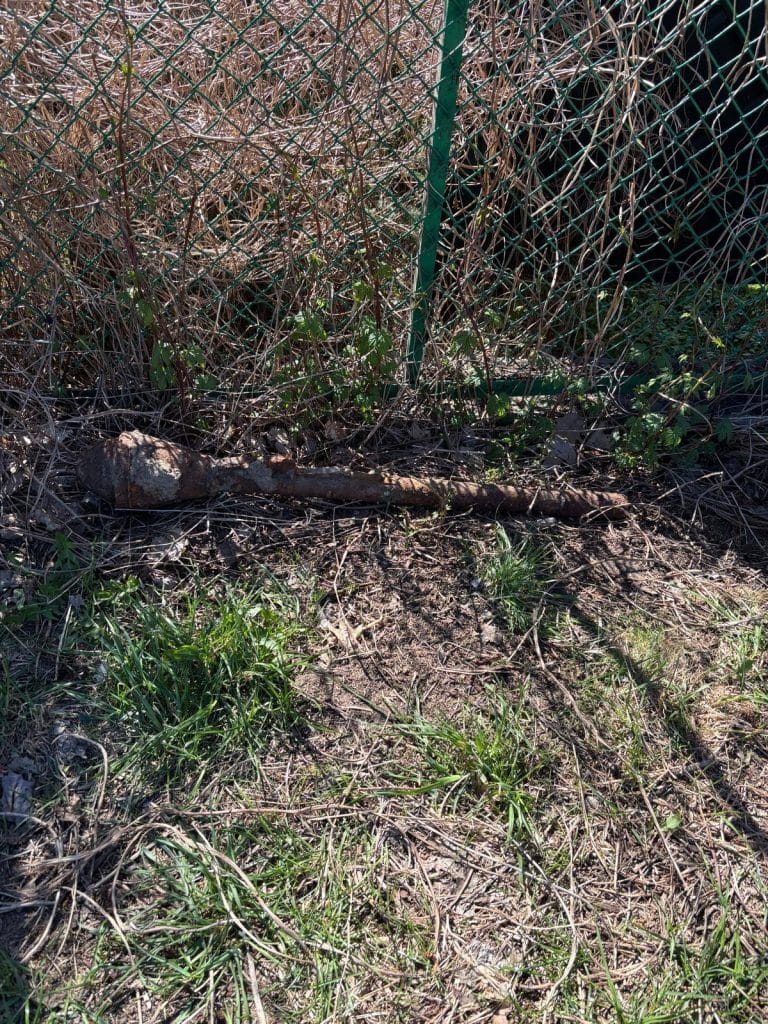Zardzewiały przedmiot na ziemi przy ogrodzeniu, teren zabezpieczony przez policję.