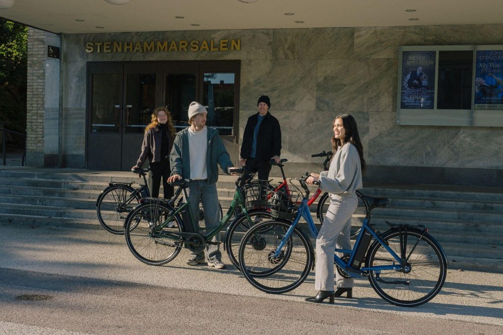 Grupa młodych ludzi z rowerami elektrycznymi przed budynkiem, uśmiechają się i rozmawiają.