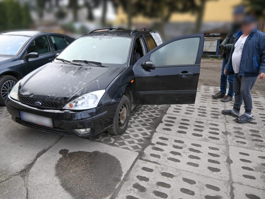 Zniszczony czarny samoch&oacute;d na parkingu, wok&oacute;ł ludzie, widoczny otwarty drzwi.