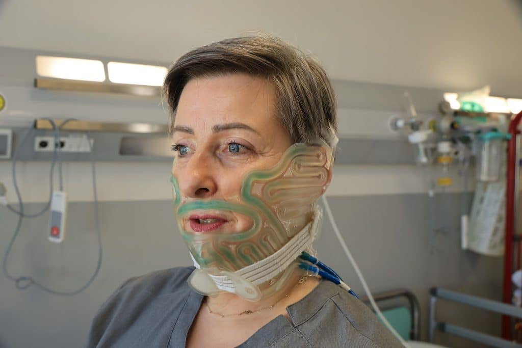 Kobieta z maską chłodzącą na twarzy podczas pobytu w szpitalu, w tle sprzęt medyczny.