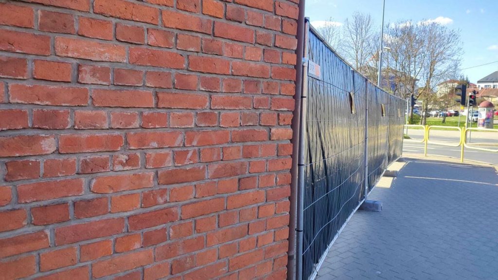 Teren wok&oacute;ł kościoła w Olsztynie zabezpieczony ogrodzeniem, widok na ceglaną ścianę i ulicę w tle.