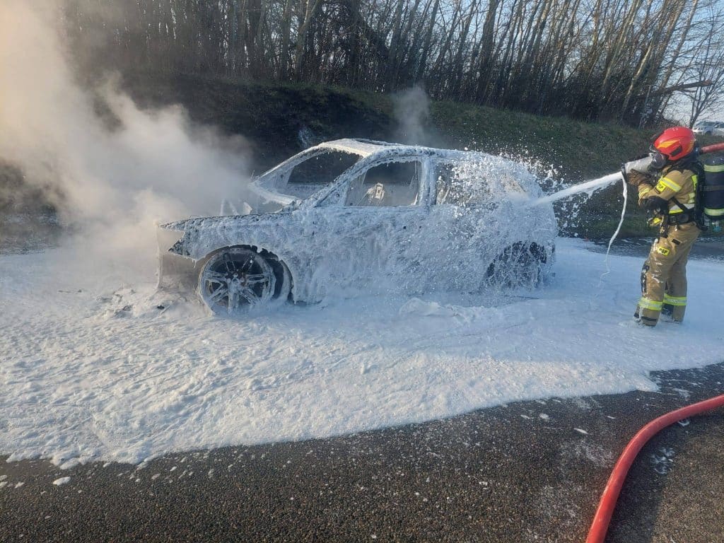 Strażak gaszący pożar samochodu na drodze ekspresowej, samoch&oacute;d pokryty pianą gaśniczą.