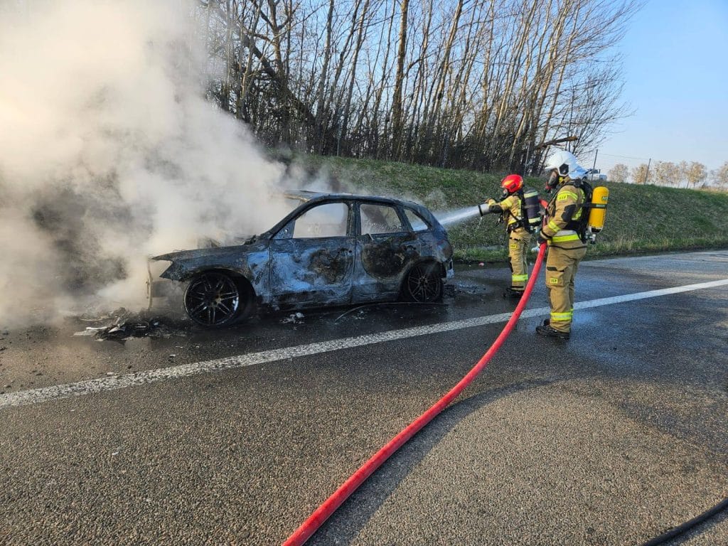 Strażacy gaszą pożar samochodu marki Audi na drodze ekspresowej S22, wok&oacute;ł widoczny dym i zniszczenia pojazdu.