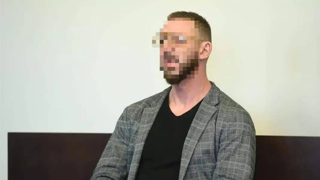 Mężczyzna z zasłoniętym twarzą, siedzący na tle białej ściany, w marynarce i czarnym T-shircie.