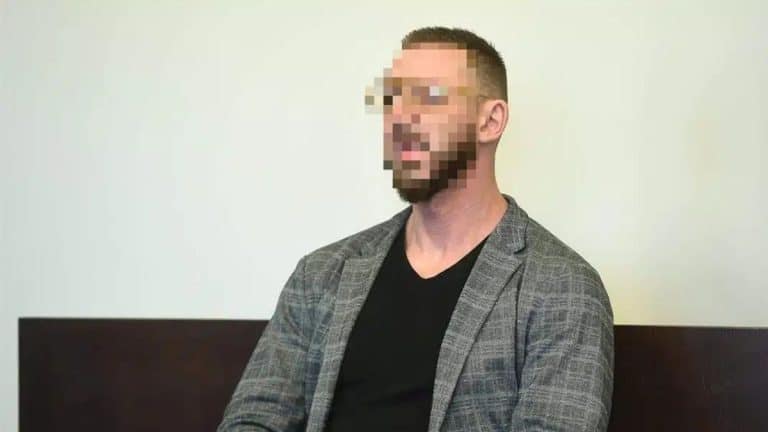 Mężczyzna z zasłoniętym twarzą, siedzący na tle białej ściany, w marynarce i czarnym T-shircie.