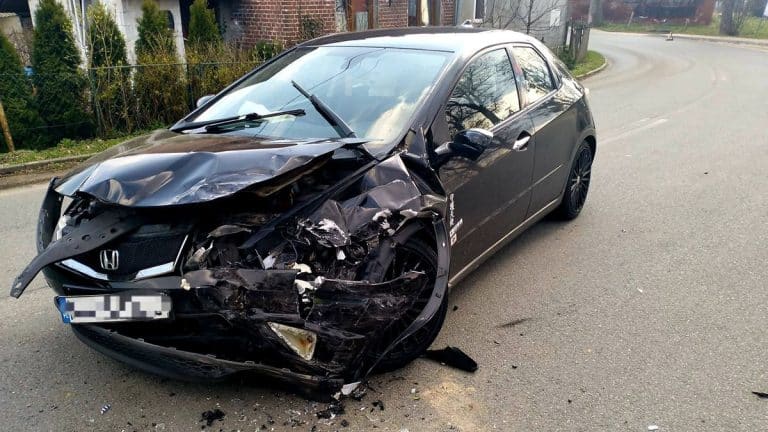 Uszkodzony czarny samoch&oacute;d po wypadku na zakręcie, widoczny rozbity prz&oacute;d i poważne uszkodzenia.