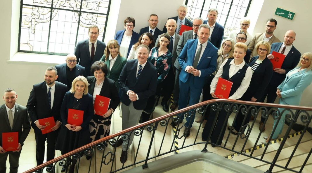 Grupa osób na spotkaniu w budynku z dużymi oknami i ozdobnym żelaznym balustradą.