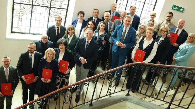 Grupa osób na spotkaniu w budynku z dużymi oknami i ozdobnym żelaznym balustradą.