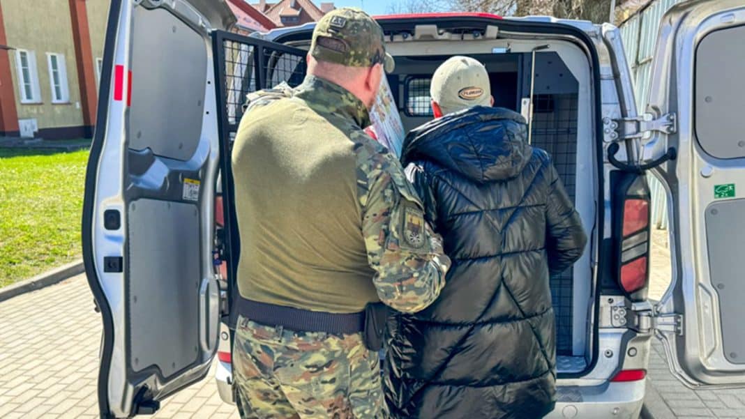 Strażnik graniczny odprowadza cudzoziemca do pojazdu na tle budynku.