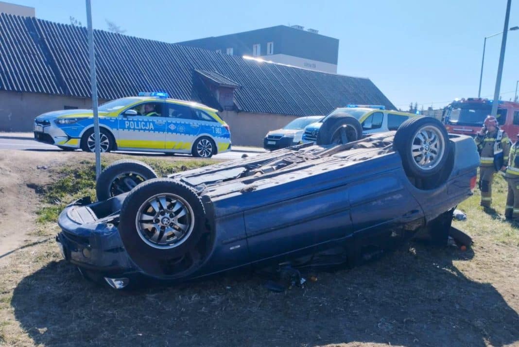 Rozbity samoch&oacute;d BMW odwr&oacute;cony do g&oacute;ry kołami na poboczu, w tle radiowozy i straż pożarna.