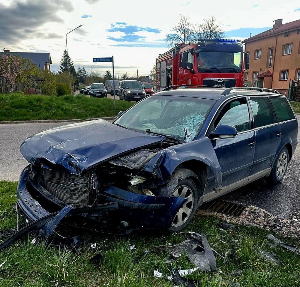 Uszkodzony samoch&oacute;d na poboczu po kolizji, w tle w&oacute;z strażacki i inne pojazdy.
