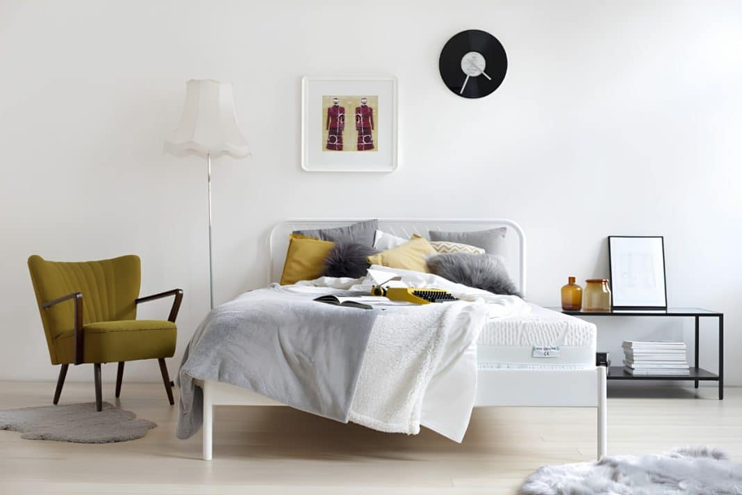 Sypialnia z ł&oacute;żkiem, na kt&oacute;rym leży pościel i poduszki, obok stolik z dekoracjami, na ścianie zegar i obraz.