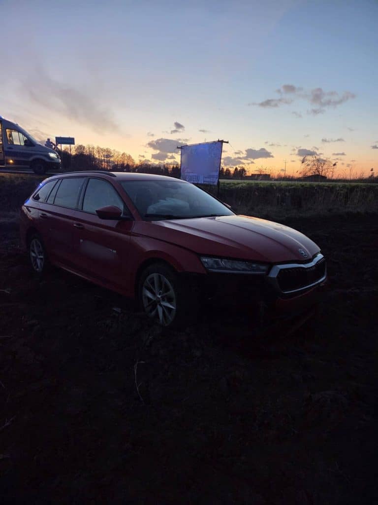 Czerwony samoch&oacute;d na polu po wypadku, w tle zach&oacute;d słońca i pojazdy na drodze.