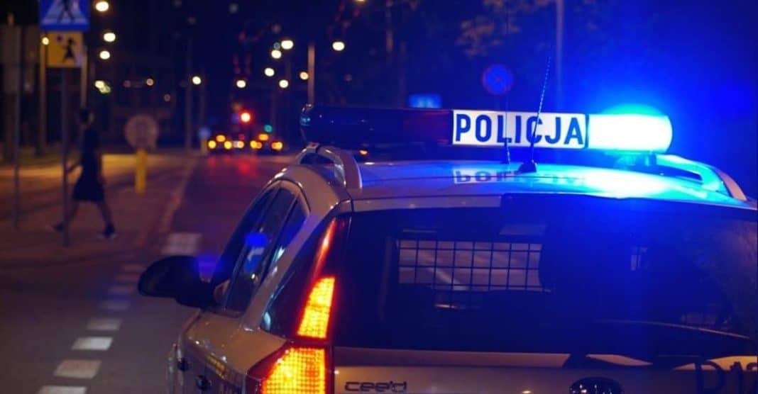 Policja na drodze w nocy, w tle widoczne osoby i oświetlenie uliczne.