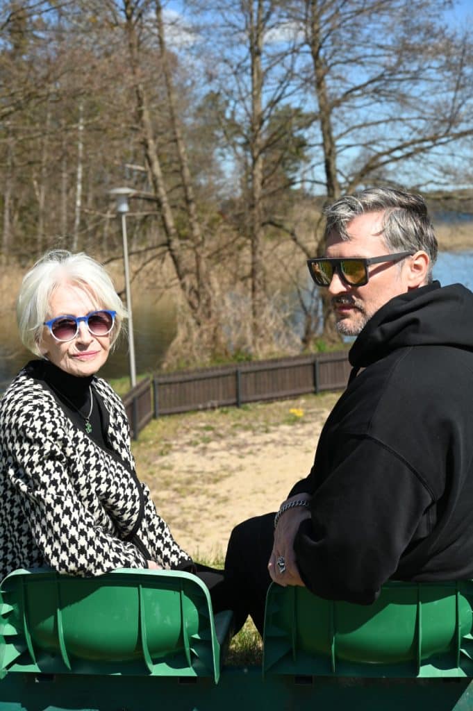 Dwoje ludzi siedzi na ławce nad jeziorem, w tle drzewa i woda, w słoneczny dzień.